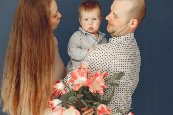 Cheerful long haired woman with bouquet of flowers embracing husband and little son while father holding boy against blue background